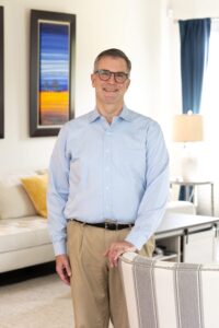 Against a background that includes a white sofa with colorful artwork above it, Johns Hopkins School of Education alum Jonathan Nateghi-Asli stands wearing tan pants, blue shirt, and glasses