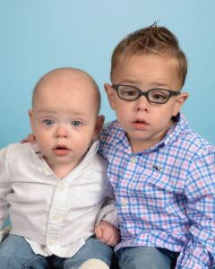 Caleb Kramer is sitting with his bother Brayden. Caleb is wearing a white collared shirt and jeans. Brayden is wearing a plaid shirt, jeans, and glasses.