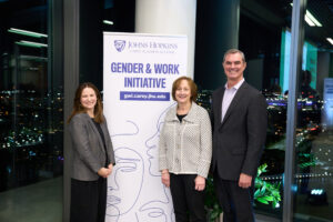 Jenny Morgan is standing with Gender and Work Initiative co-directors Colleen Stuart, PhD, (right) and David Smith, PhD (left) and the initiative launch event. They are standing in front of the Gender and Work Initiative backdrop. They all are wearing business attire. 