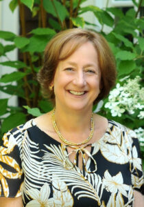 Jenny Morgan is smiling and standing outside. She's wearing a floral blouse with a gold necklace.