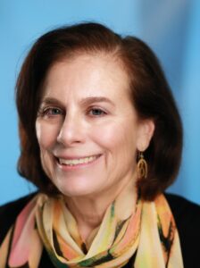 Julie Reinganum is smiling and sitting in front of a blue backdrop. She's wearing a black sweater with a colorful scarf and gold-colored earrings.