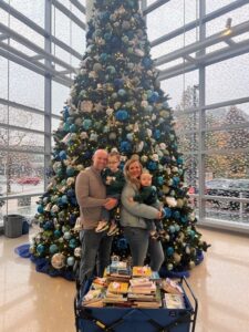 Thomas and Emily Kramer are standing with their two boys in front of a decorated Christmas tree at the Johns Hopkins Children Center. Thomas is holding Brayden and Emily is holding Caleb with a wagon full of children's books.