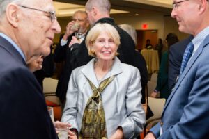 Sheila Wharam wears a light gray suit with a wide gold scarf as she smiles and stands between two men in suits