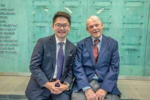 Sitting on a bench with a glass wall behind them are Wilmer Eye Institute physician T.Y. Liu in a dark blue suit, striped tie and glasses and donor James Gills in gray pants, red tie and blue jacket