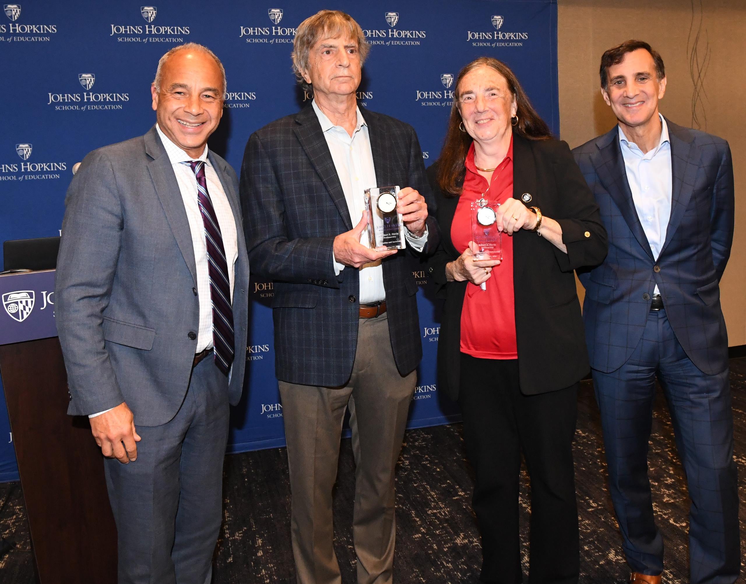 Christopher Morphew, PhD, dean of the School of Education, Steven Ross, PhD, Nancy Madden, PhD, and President Ron Daniel are standing in front of a Johns Hopkins School of Education backdrop. They are all smiling. Steven Ross and Nancy Madden are each holding on to a plaque.