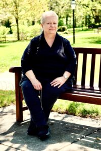Cathy Lonas is sitting and smiling on a wooden bench at a park. She's wearing a black blouse and dress pants. There are bloomed trees and green grass behind her.