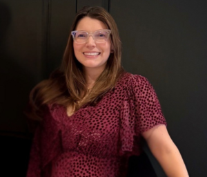 Lee-Lee Ellis is smiling and standing in front of a dark-wooded door frame. She's wearing clear glasses and a burgundy-colored dress.