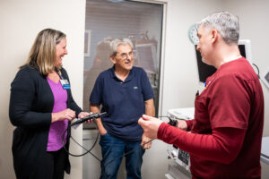 Jennifer Moody, Tony Roussos, and Denis Kotsoev are in conversation and smiling at each other. Moody and Kotsoev are holding the black fiberoptic endoscopic scope and demonstrating how it works.