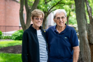 Laura and Tony are standing and smiling outside. Laura is wearing a black and white striped sweater, with a black jacket, and Tony is wearing a blue polo. They are standing in front of bloomed trees.