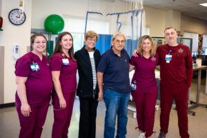 Laura and Tony Roussos are standing with Tony's four therapists in the comprehensive inpatient rehabilitation unit. The Roussos are in the middle of the group. Everyone is smiling.