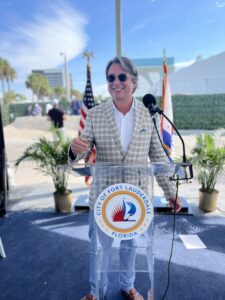 Jon Kling is standing behind a clear podium smiling in a beachy area of Fort Lauderdale, Florida. He's wearing sunglasses, a white collared shirt, a tan-colored plaid jacket, and light blue pants. There are small palm trees behind him.