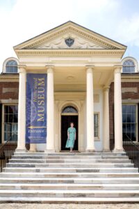 Eileen Perkins is standing in between two pillars on the steps of Homewood House. She's wearing a green flowy dress with a white flower pattern.