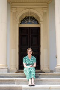 Eileen Perkins is sitting on Homewood Museum's front steps. She's wearing a long flowy green dress with a pattern of white flowers. The arch doorway of the house is behind her.