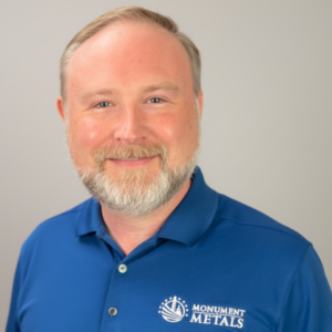 Jonathan Swyers is smiling in front of a white backdrop. He's wearing a blue polo which features Monument Metals' logo.