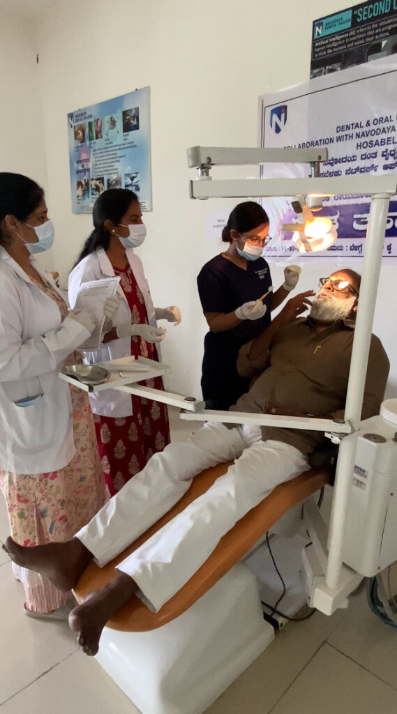 Anupama John, wearing blue scrubs, and two fellow dentists, wearing white coats and dresses, perform a dental screening on an individual at Navodaya Dental College and Hospital in India.