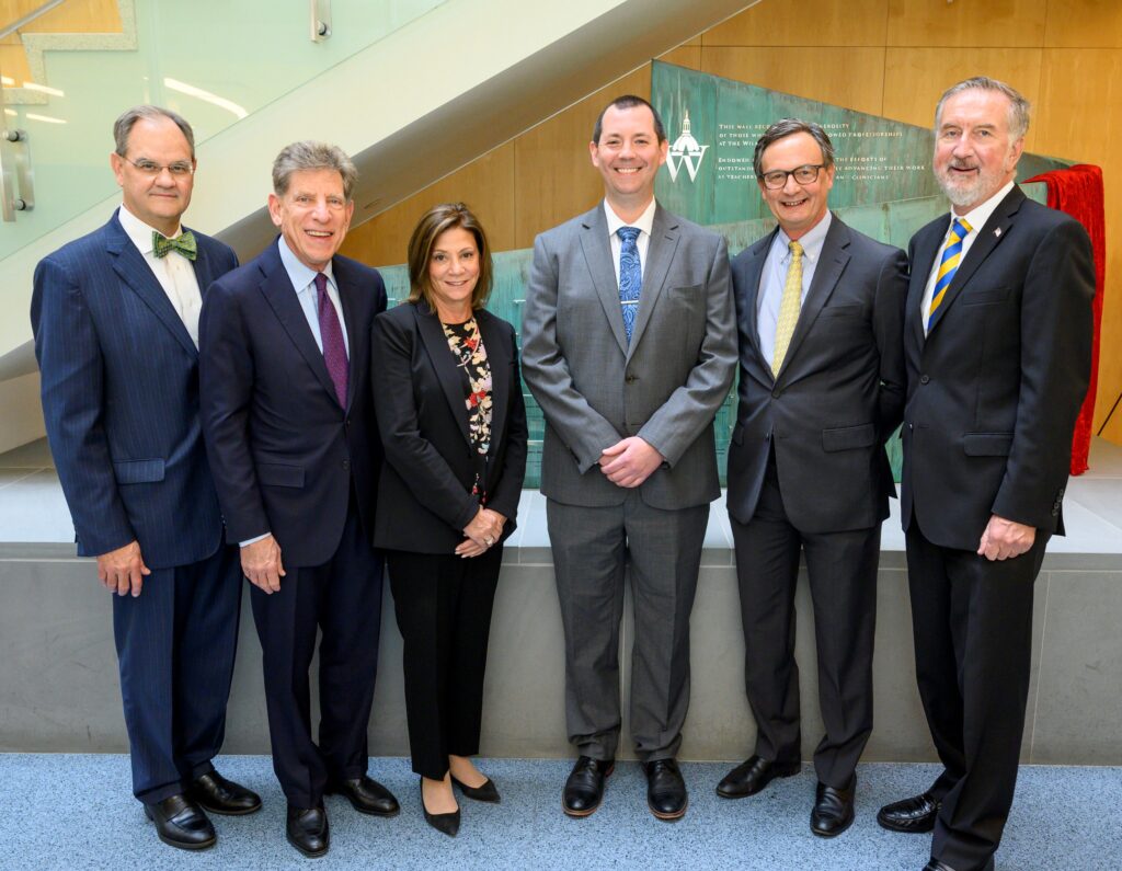A group of six people, four men and one woman, all affiliated with Wilmer are standing and smiling. Everyone is wearing business attire suits. 