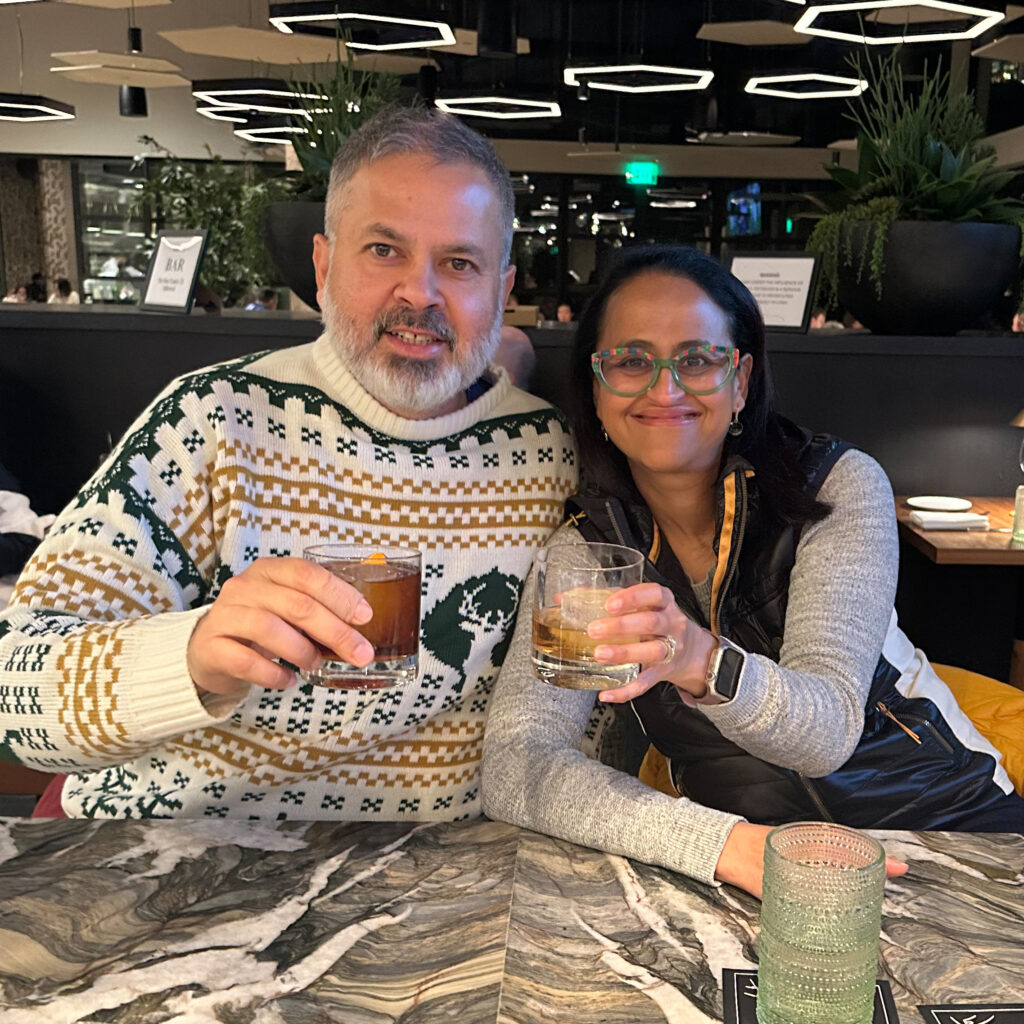 Girish and Himangi Rishi sit together at a resturant cheering their drinks and smiling.